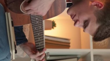 Vertical video. Male songwriter. Home leisure. Creative life. Inspired man playing guitar composing music sitting couch light room interior.