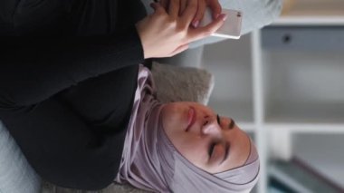 Vertical video. Mobile chat. Gadget weekend. Online home leisure. Happy relaxed woman in hijab using phone social media app on couch at living room.