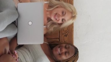 Vertical video. Girls online weekend. Digital leisure. Sleepover morning. Two joyful multicultural female friends watching movie together on laptop.