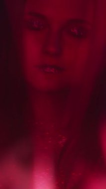 Vertical video. Color light face. Nightclub beauty. Fashion look. Blur neon red woman with shimmering glitter artistic makeup metallic eyeshadow lips dancing on dark.