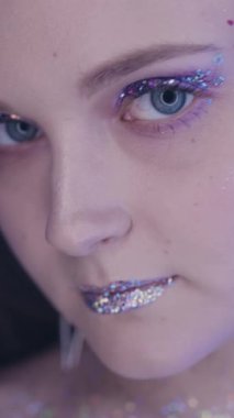 Vertical video. Glitter face. Artistic makeup. Nightclub look. Closeup of glamour woman with sparkling skin metallic eyeshadow lips in blue neon color light.