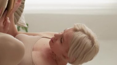 Vertical video. Exciting book. Curious woman. Interesting story. Inspired middle-aged lady catching of reading sitting sofa in light home interior.