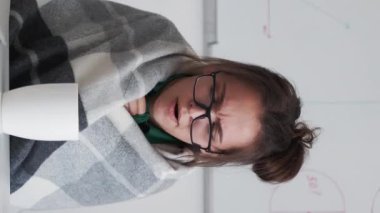 Vertical video. Sick business woman. Online meeting. Remote conference. Unwell female office worker in blanket with flu sneezing feeling cold working from home drinking tea.