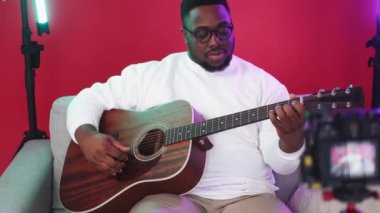 Music broadcasting. Video blog. Internet streaming. Talented man playing acoustic guitar recording social media content on camera sitting on couch.