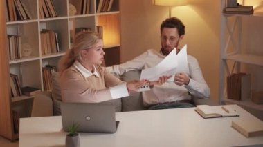 Bad work. Displeased woman. Colleagues quarrel. Angry female manager showing wrong documents to relaxed careless man sitting sofa drinking coffee in light room interior.