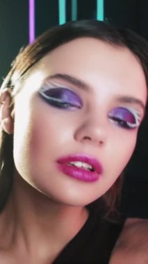 Vertical video. Disco makeup. Fashion girl. Retro 90s beauty. Confident teen model face with blue color eyeshadow pink lips artistic look posing in neon light on black.