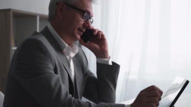 Business phone call. Mobile cooperation. Information technology. Successful confident middle-aged man talking working on tablet at light office interior with free space.
