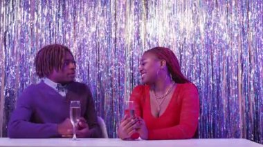 Bad date. Quarreling couple. Holiday meeting. Unhappy displeased black woman trying to avoid obsessive man sitting desk with champagne on shimmering cascade curtain background.