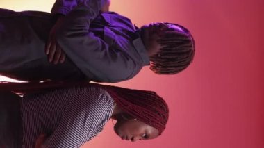 Vertical video. Offended couple. Neon light portrait. Aggrieved black woman and man staying back each other keep silence on purple peach pink background.