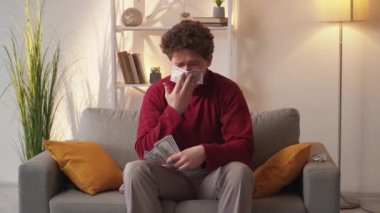 Useless money. Bankrupt man. Depressed feelings. Desperate guy crying cleaning tears blowing nose with cash dollars sitting sofa in light room interior.