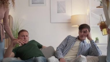 Uninterested leisure. Bored male friends. Drink inspiration. Happy women proposing to men beer to have rest at home on sofa light room interior.