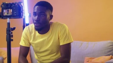 Vlog film. Video interview. Live streaming. Confident influencer man on couch speaking recording content on camera in lighting equipped yellow studio with free space.