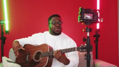 Guitar class video broadcasting. Musician man teaching playing acoustic instrument recording music tutorial on camera in lighting equipped red studio.