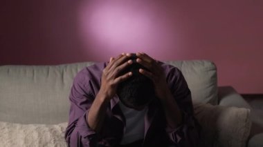 Stress headache. Anxiety attack. Nervous breakdown. Depressed frustrated man suffering from pain clutching head crying on couch on purple free space.