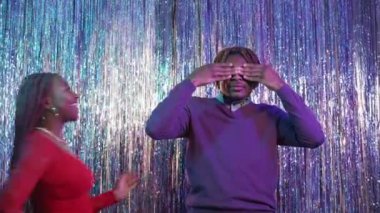 Festive gift. Beloved couple. Christmas surprise. Amazed black man closed eyes getting present box from excited woman posing shimmering cascade curtain background.