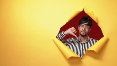 Dislike gesture. Negative feedback. Bad idea. Disappointed man showing thumb down in yellow breakthrough hole ripped paper wall on advertising copy space background.