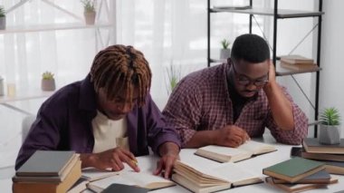 Home education. Male student. Preparing exam. Exhausted black men sitting desk studying books in light room interior.