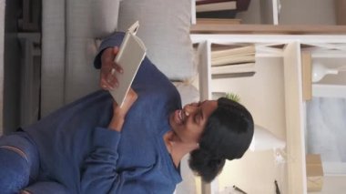 Vertical video. Reading joy. Happy woman. Home relax. Joyful lady enjoying book sitting floor light living room interior.