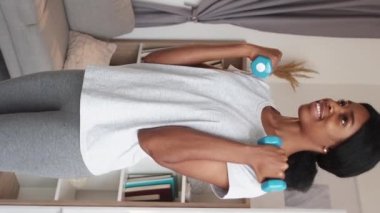 Vertical video. Dumbbells exercise. Happy woman. Sporty leisure. Cheerful athletic slim lady in sportswear practicing muscle workout indoors at living room.