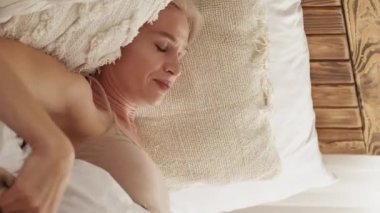 Vertical video. Bad sleep. Displesed woman. Uncomfortable bed. Angry lady spinning on inconvenient mattress and pillows impossible to rest light home interior.