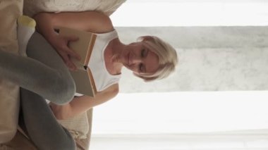 Vertical video. Home leisure. Reading woman. Enjoying weekend. Happy middle-aged lady sitting comfortable couch with interesting book in light room interior.