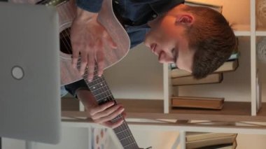 Vertical video. Online training. Male teacher. Guitar practice. Inspired man teaching playing on string instrument on laptop sitting light room interior.