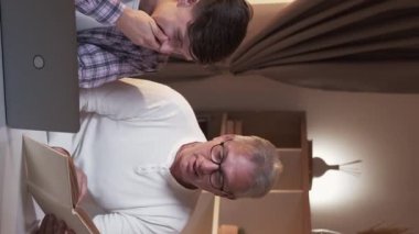 Vertical video. Father son study. Parent teaching. Home education. Two joyful men laughing enjoying reading funny book together at workplace.