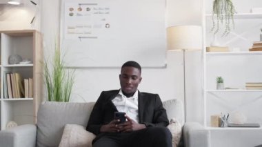Mobile business. Millennial work. Phone research. Pensive man employee browsing smartphone internet chatting online on office couch copy space.