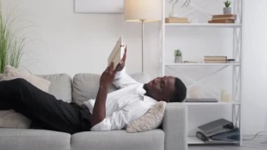 Book relaxing. Office leisure. Reading recreation. Happy peaceful man resting on cozy couch enjoying break in light room interior.