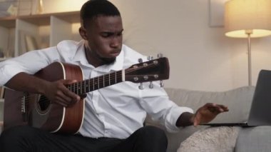 Guitar course. Hard education. Displeased man. Frustrated male guitarist learning string instrument on laptop video lesson in light room interior.