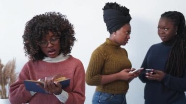 Blaming people. Smart woman. Friends gossip. Female friends with smartphones discussing clever inspired student reading book in light room interior.