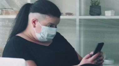 Salgın muhabbeti. Uzaktan iletişim. Karantina karantinası. Polyethylene 'de internetten mesaj atan koruyucu maskeli şişman bir kadın ev içi polietilen kaplı..