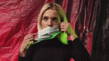 Environmental pollution. Allergic disease. Ecology problem. Woman behind transparent net putting protective mask on red green neon light wrinkled plastic film background.