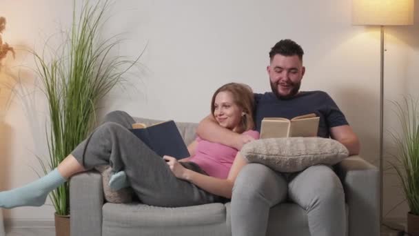 ホームレジャー リラックスしたカップル 家族関係だ ポジティブな男と女読書本一緒に座っています上のソファで光の部屋のインテリア — ストック動画