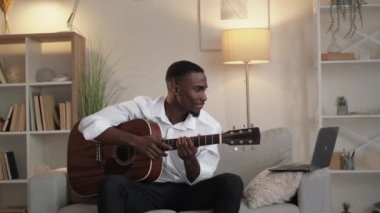 Online lesson. Music school. Remote education. Happy man learning guitar looking video on laptop sitting sofa light home interior.