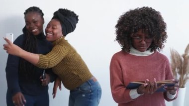 Outcast person. Social life. Friends jealousy. Happy women making selfie together while upset female with book staring them in light room interior.