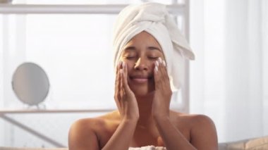 Skin moisturizing. Facial skincare. Beauty routine. Satisfied relaxed smiling woman applying nourishing face cream after morning shower in light bathroom with free space.