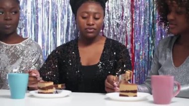 Party celebration. Female talk. Sweet treats. Happy black women eating festive cake sitting desk discussing news on cascade curtain background.