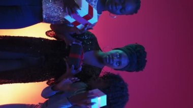 Vertical video. Holiday present. Curious women. Neon light people. Festive female friends shaking gift boxes on blue pink background.