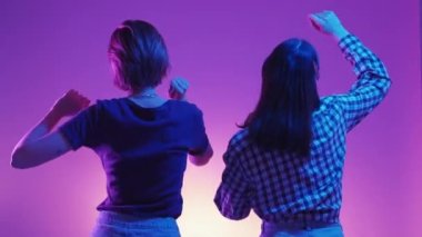 Dance party. Female friends. Neon light people. Unrecognizable two happy women moving on music together posing green violet background back view.