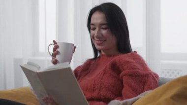 Home reading. Relaxed woman. Weekend leisure. Happy female sitting sofa with interesting book cup of coffee in light room interior.