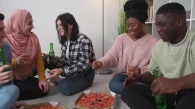 Pizza party. Happy friends. Students leisure. Funky international men and women enjoying communication with fast food and beer in light room interior.