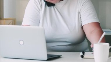 Executive work. Remote consulting. Multitasking employee. Confident overweight woman using laptop talking on phone taking notes at light home office workplace.