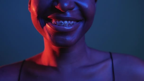 笑顔の美しさ 歯列矯正ケア 歯科治療 暗い上に隔離されたネオン赤青の色の光で金属歯のブレースで認識できない幸せな陽気な女性の顔 — ストック動画