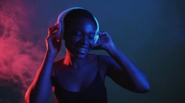 Cyber music. Sound technology. Electronic techno. Neon light blue red inspired relaxed woman enjoying listening rhythm in wireless headphones in color smoke isolated on dark night.