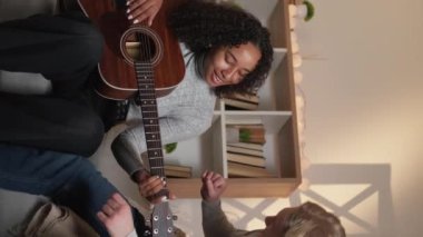 Vertical video. Guitar playing. Home lesson. Beloved couple. Inspired man showing woman accords on acoustic instrument sitting light room interior.