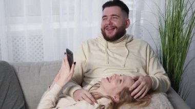 Mobile memories. Happy couple. Home leisure. Laughing man and woman looking smartphone resting sofa together in light room interior.