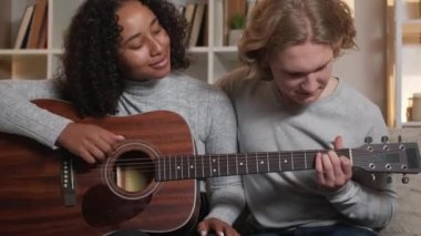 Music lesson. Beloved couple. Home leisure. Happy man and woman playing guitar together sitting sofa in light room interior.