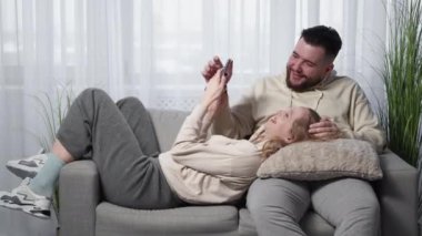 Mobile shopping. Happy couple. Family leisure. Laughing man and woman choosing product smartphone resting sofa together in light room interior.