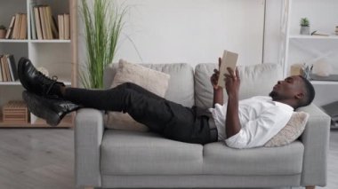 Work break. Office relax. Home leisure. Tired yawning man reading book napping on cozy sofa in light room interior.